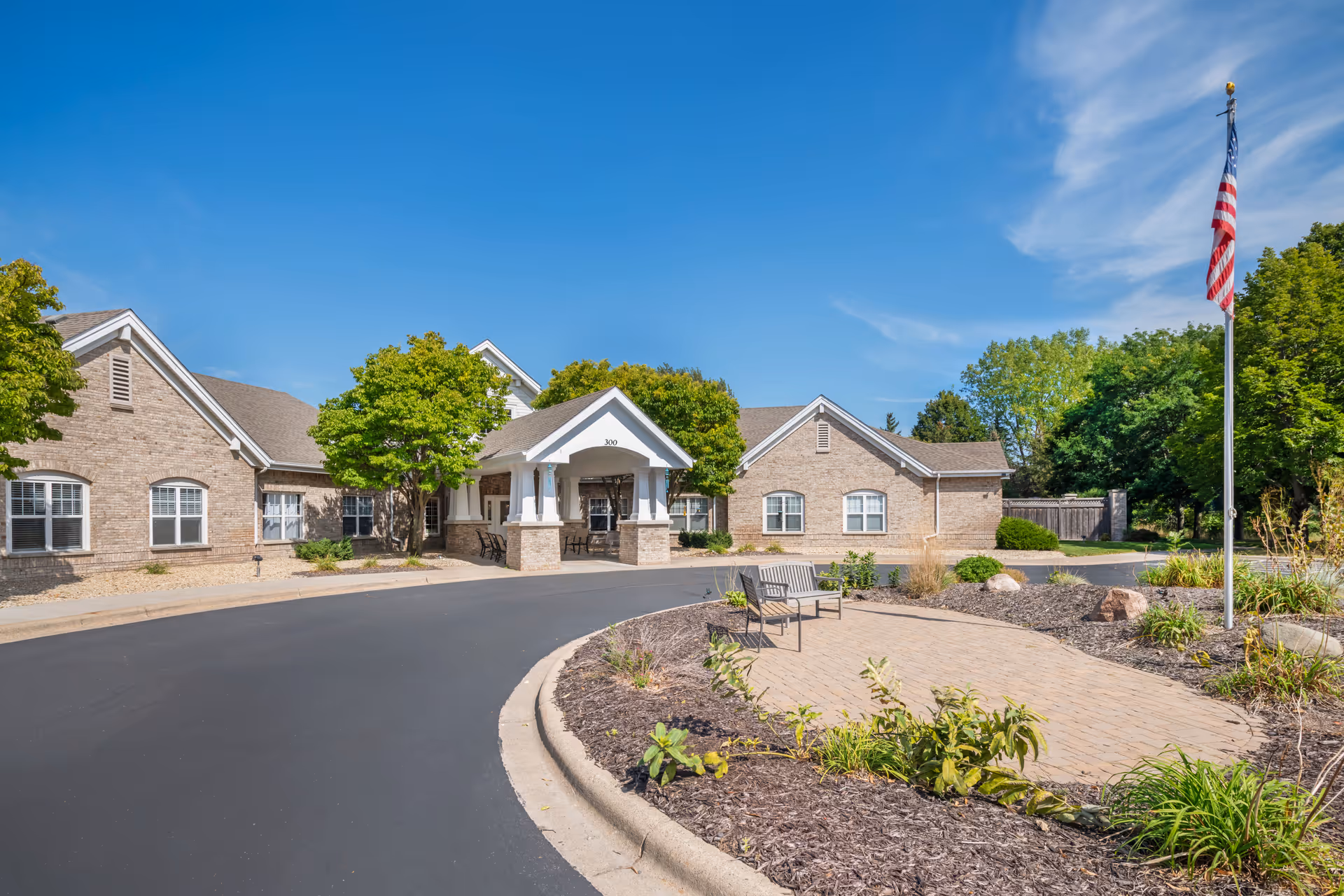 Exterior view of Brookdale North Oaks facility showing a brick building with a covered entrance, a curved driveway, landscaped garden beds with plants and mulch, two benches on a paved area, and an American flag on a flagpole under a clear blue sky.