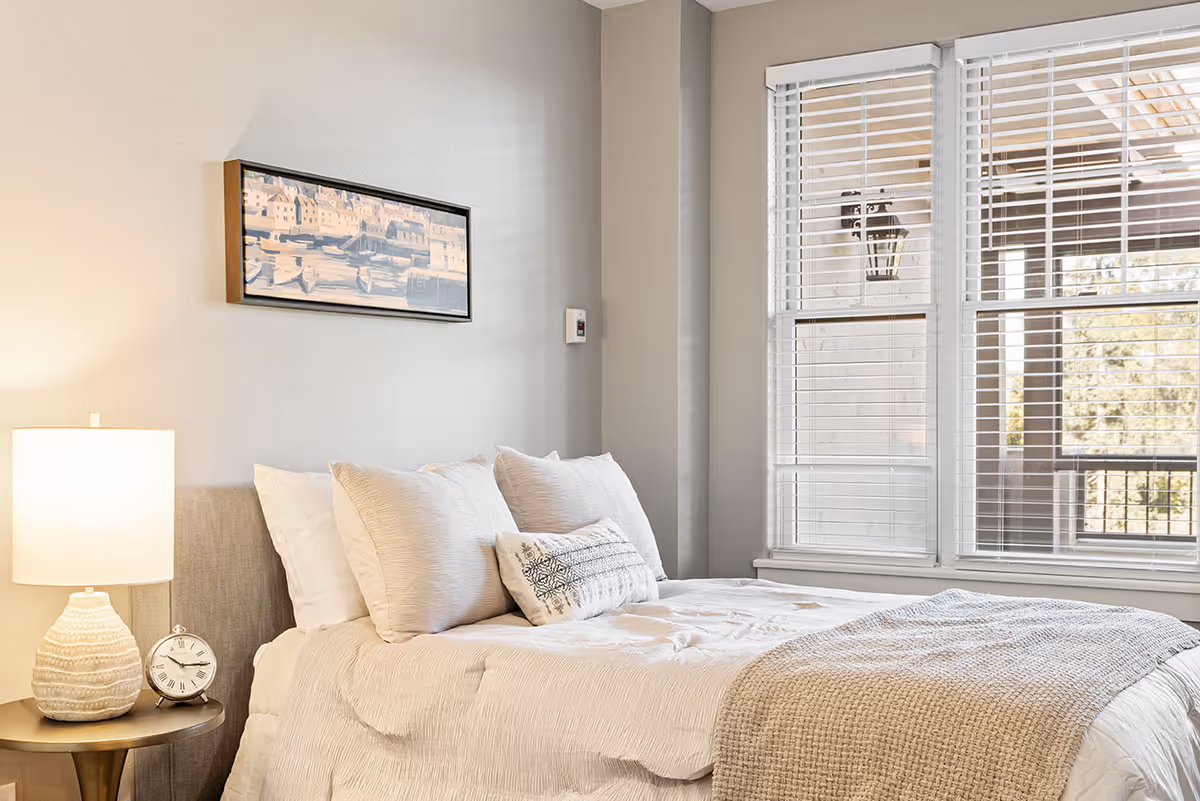 Bright bedroom with a neatly made bed, bedside table and lamp, wall art, and large windows with blinds.