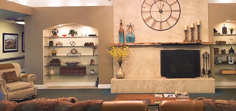 A cozy living room area featuring a large stone fireplace with a decorative clock above it. On either side of the fireplace are built-in shelves displaying various decorative items such as vases, books, and small sculptures. Comfortable armchairs and sofas are arranged around a wooden coffee table with magazines on it.