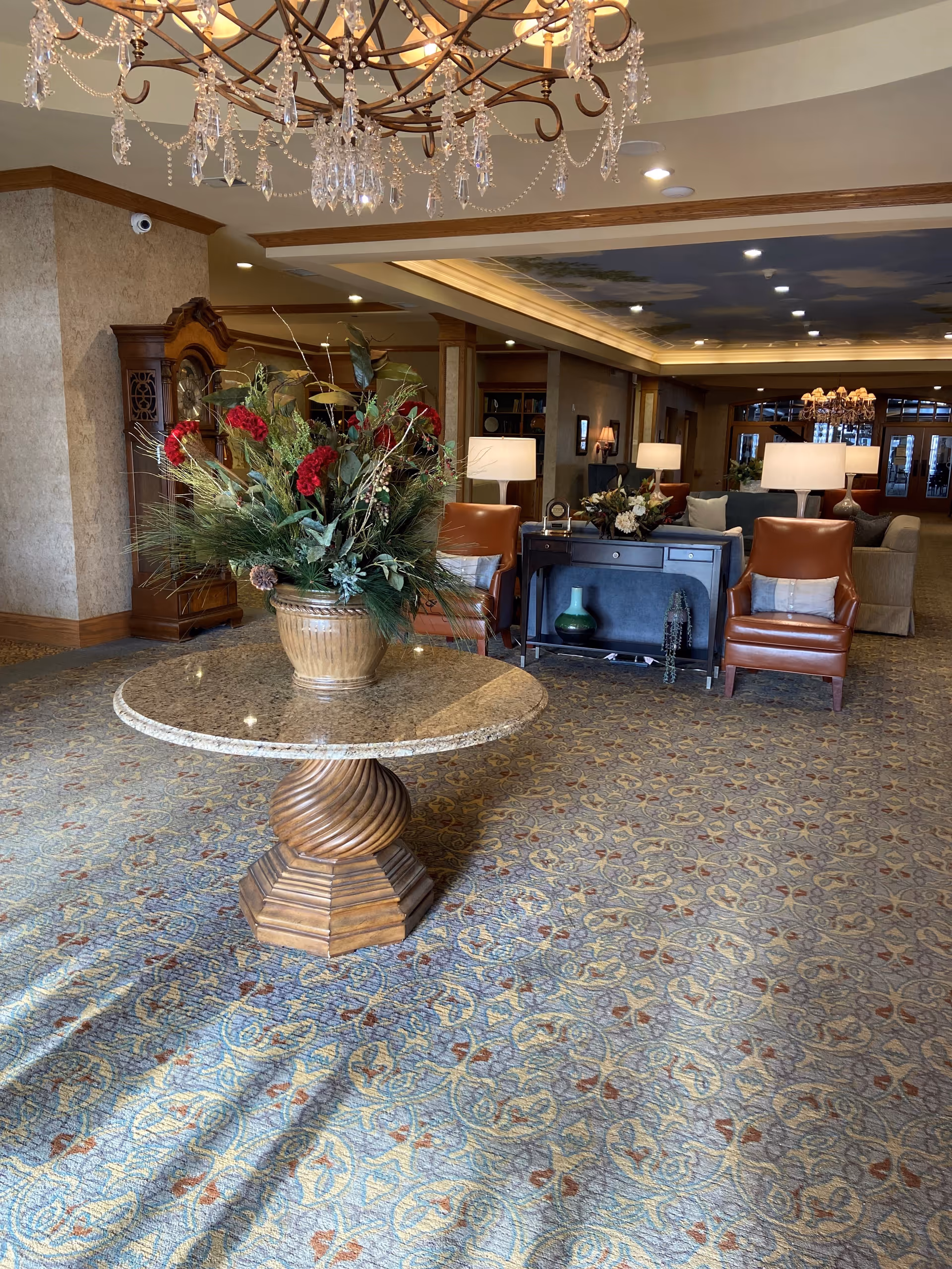 A spacious and elegantly decorated senior living facility common area with a round wooden table topped with a large floral arrangement in the foreground. The room features patterned carpet, leather armchairs, a console table with decorative items, multiple lamps, and a chandelier hanging from the ceiling. The ceiling has a painted sky design, and there is a grandfather clock against the wall.