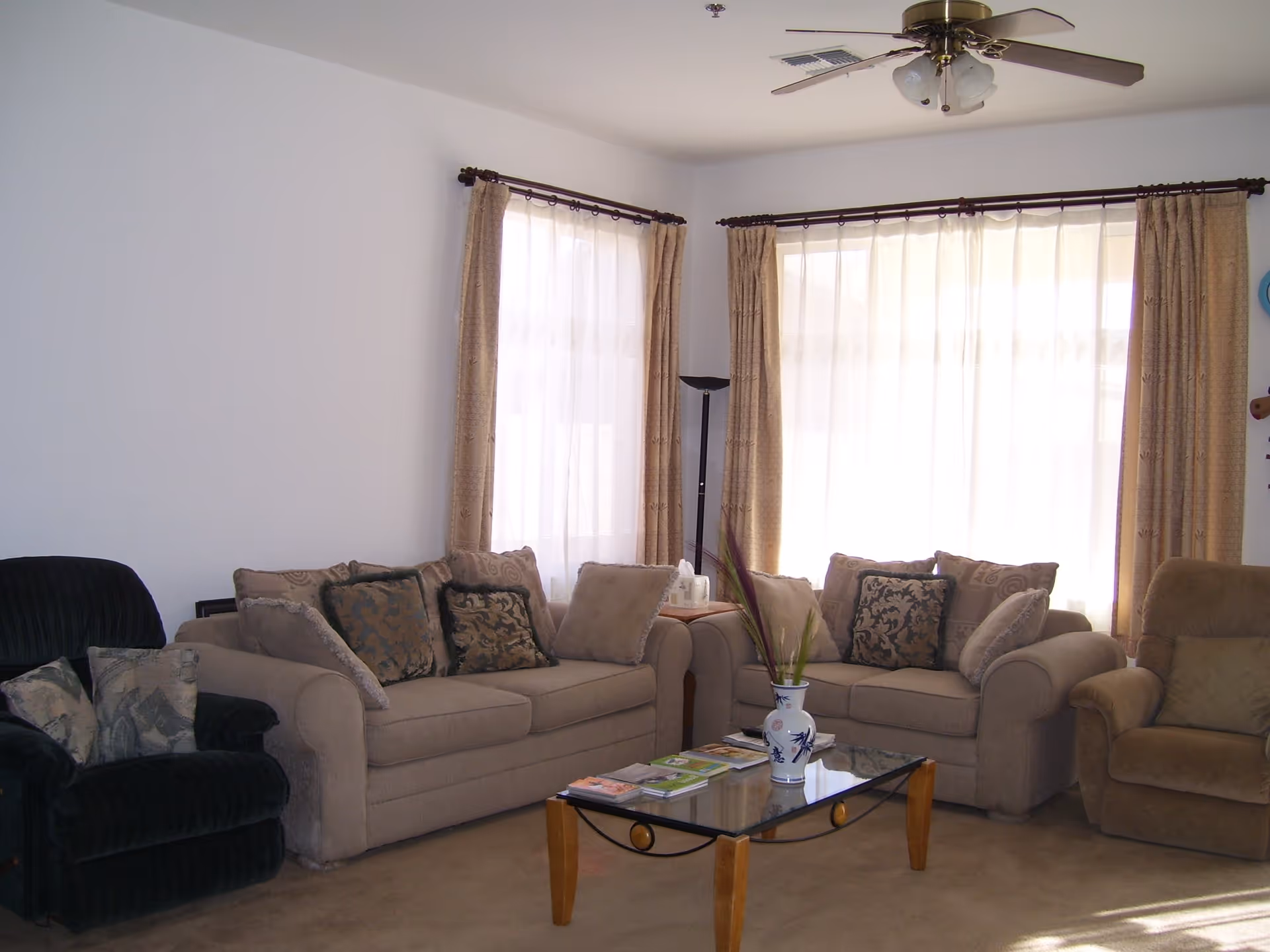 A living room with two beige sofas and two armchairs arranged around a glass coffee table. The coffee table has a decorative vase with plants and some magazines on it. The room has two large windows with sheer white curtains and beige drapes. A ceiling fan with lights is mounted on the white ceiling.