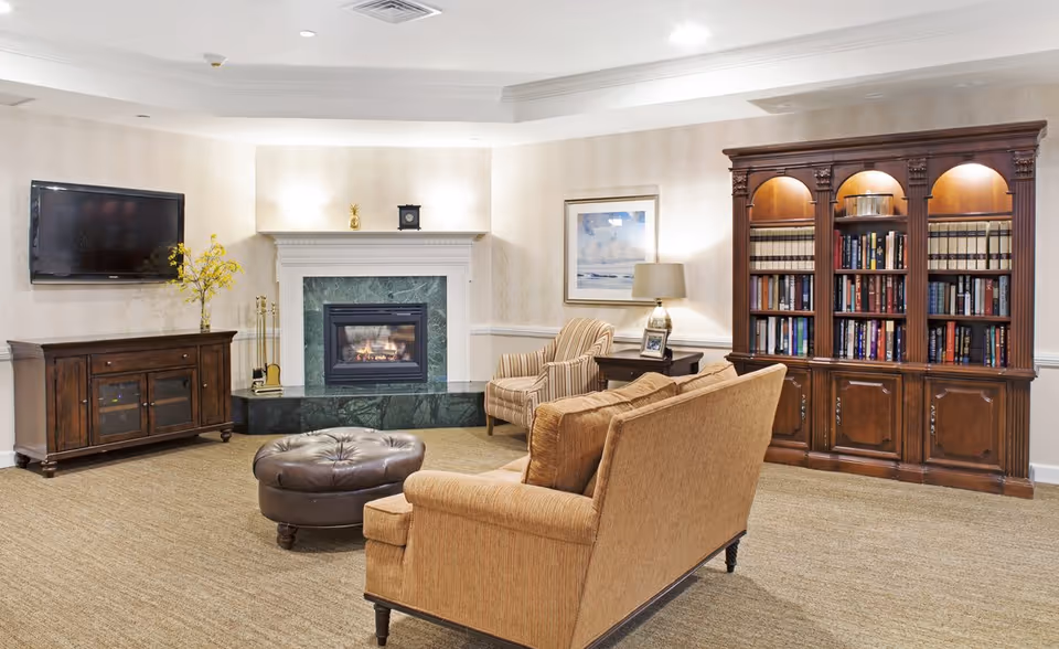 Cozy senior living lounge with sofa, armchairs, a fireplace, wall-mounted TV, and a large wooden bookcase.