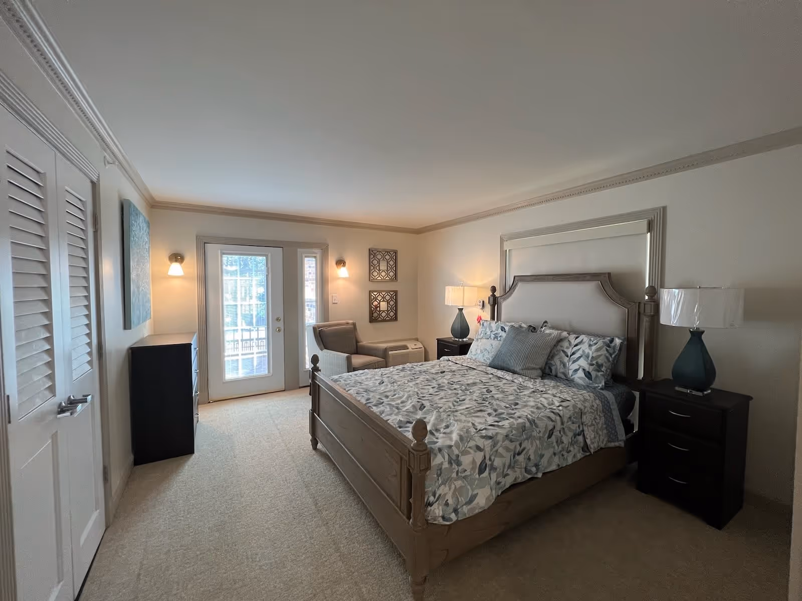 A well-lit bedroom with a large bed featuring a floral patterned bedspread and multiple pillows. There are two dark wood nightstands on either side of the bed, each with a lamp. A comfortable armchair is placed near a set of glass doors leading outside. The room has light-colored walls and carpeted flooring, with a dresser and wall art on the left side.