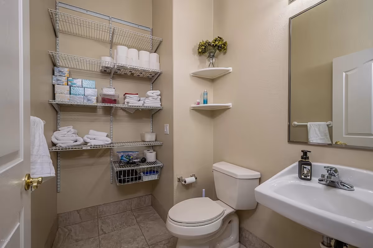 A small bathroom with a white toilet and a white sink with a soap dispenser. There is a large mirror above the sink and a towel hanging on a rack on the door. On the left wall, there are metal wire shelves stocked with toilet paper, tissues, and folded towels. Two corner shelves above the toilet hold a small vase with green foliage and some small bottles.