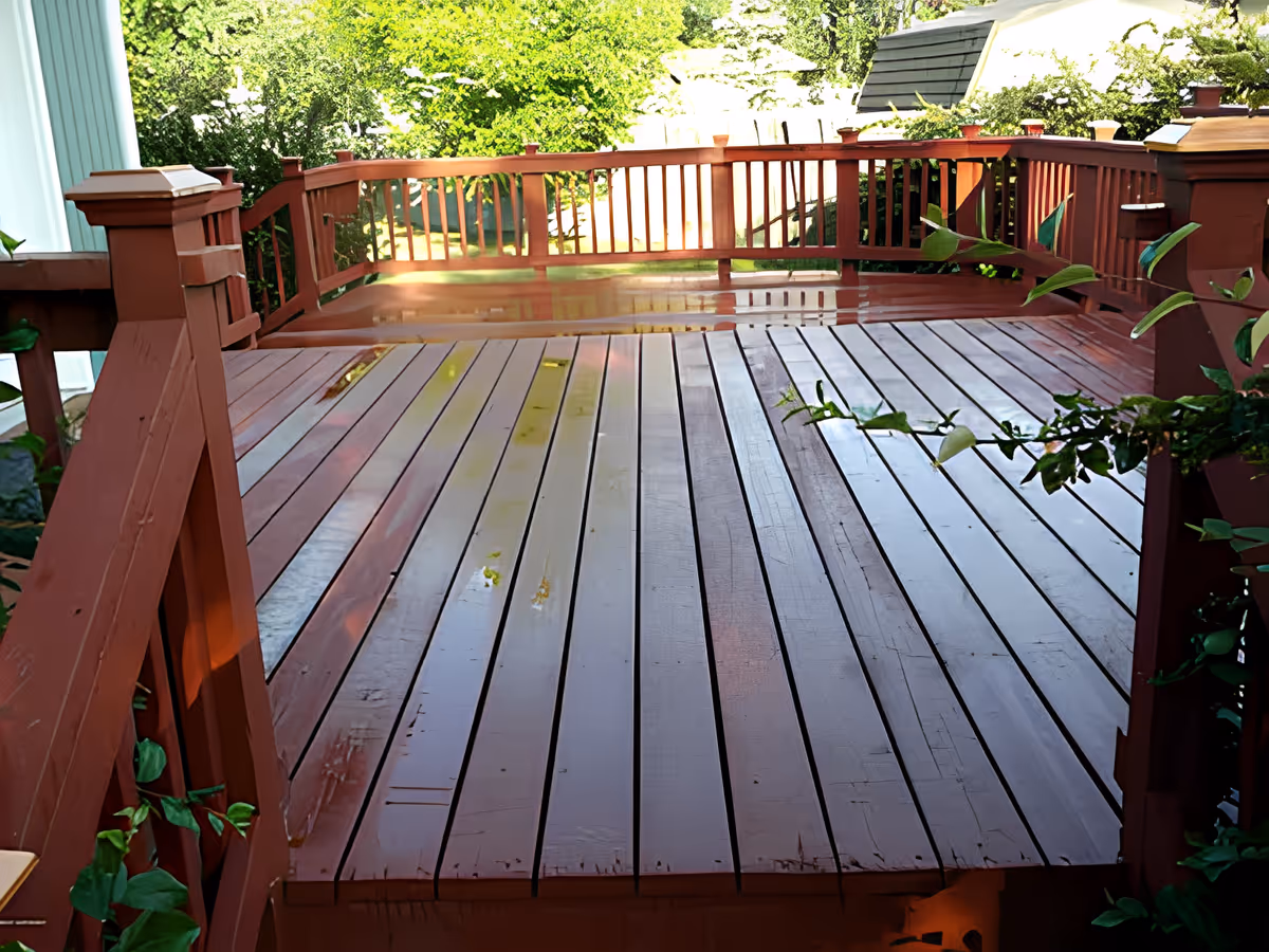 A wooden outdoor deck with reddish-brown railings and floorboards, surrounded by green foliage and trees in the background.