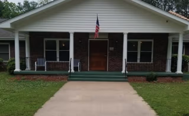 Front entrance of a single-story brick senior living building with a covered porch, white columns, an American flag, and a concrete walkway.