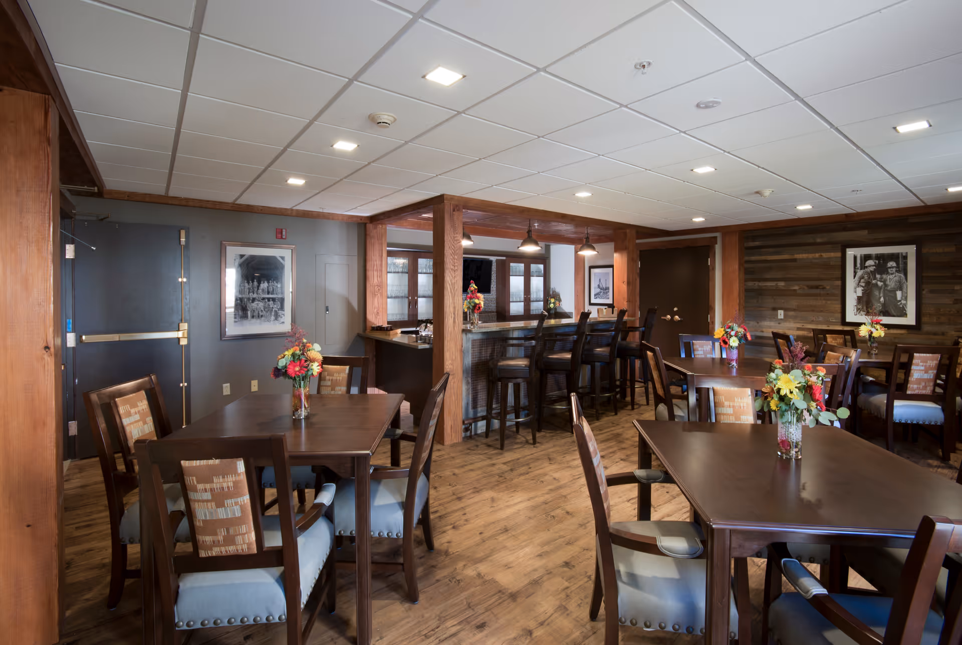 Interior view of a dining area with wooden tables and chairs, each table decorated with a vase of colorful flowers. The room features wood flooring, a bar area with high stools, and framed black and white photographs on the walls. The ceiling has recessed lighting and the overall decor has a warm, rustic feel.