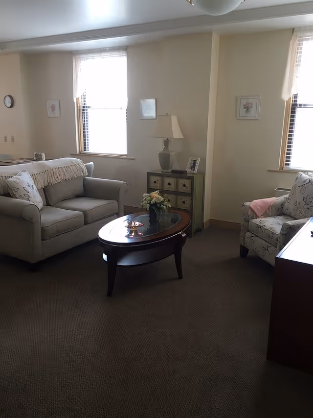 Cozy living room with a sofa, armchair, coffee table, side table with lamp, and two windows.