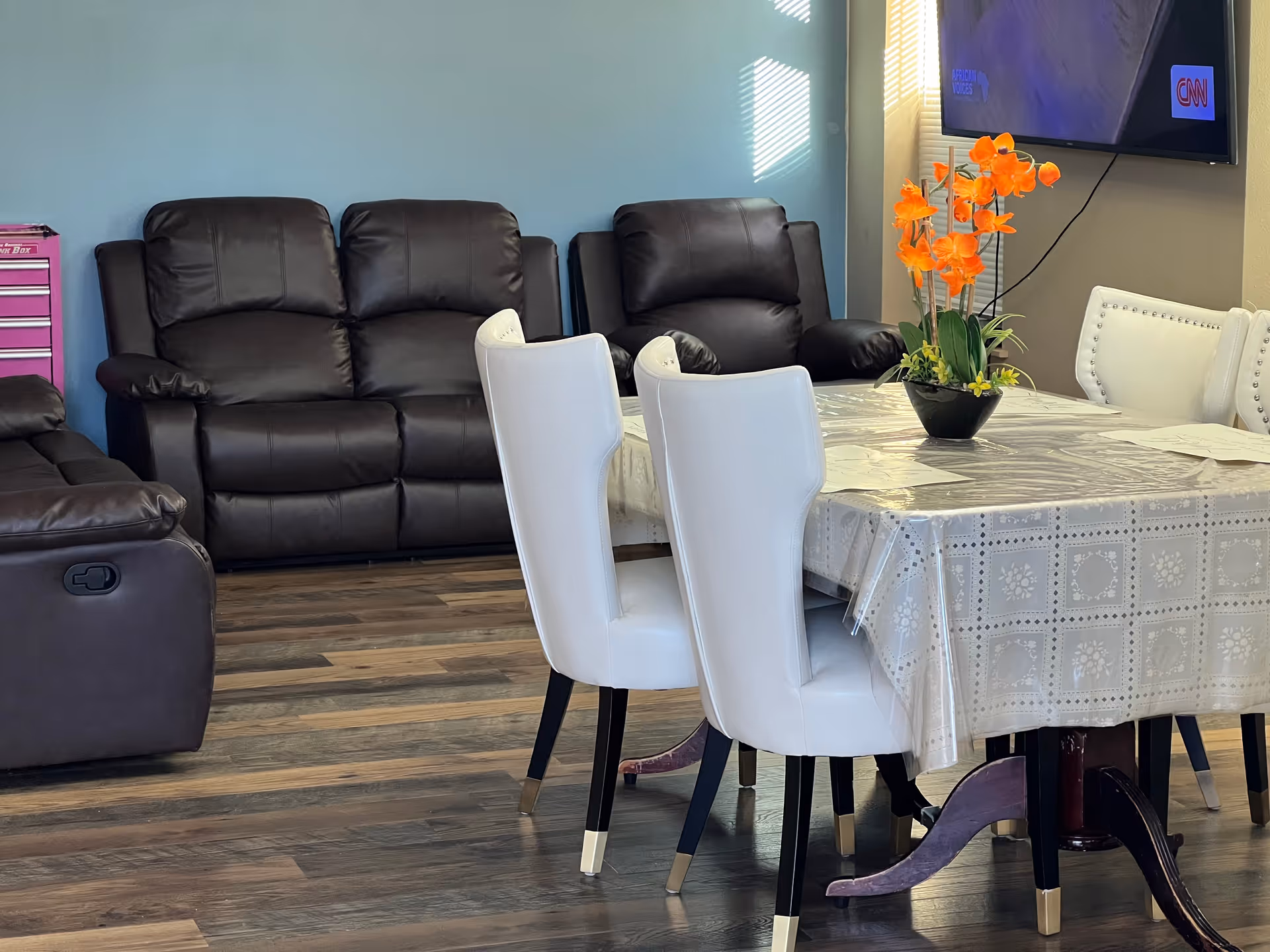 A living room and dining area with dark brown leather recliner sofas against a blue wall, a dining table covered with a patterned tablecloth and clear plastic cover, surrounded by white chairs with gold-tipped legs. An orange flower arrangement is placed on the table. A flat-screen TV is mounted on the wall in the background.