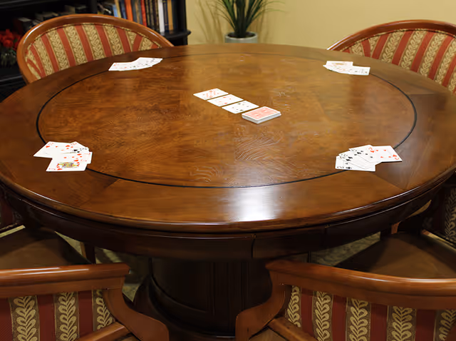 A round wooden table with playing cards spread out in four sets around the table. Four wooden chairs with red and gold patterned cushions surround the table. In the background, there is a bookshelf and a potted plant.