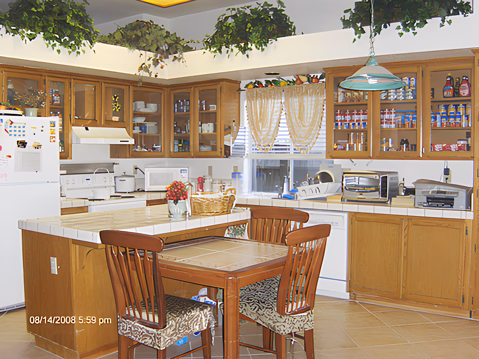 A well-lit kitchen with wooden cabinets and a white tiled countertop island. The kitchen features a white refrigerator, stove, microwave, toaster oven, and various kitchen appliances. There are four wooden chairs with patterned cushions around a small dining table attached to the island. The window has sheer curtains, and there are decorative plants on top of the cabinets.