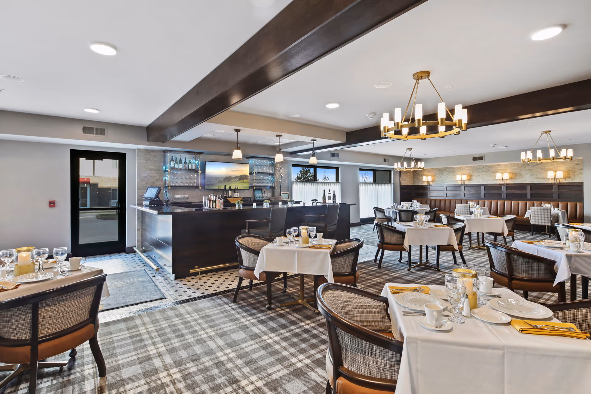 A bright and elegant dining room with tables covered in white tablecloths, set with plates, glasses, and yellow napkins. The room features plaid carpet flooring, comfortable cushioned chairs, and a dark wood bar area with pendant lights and a TV mounted on the wall. Large windows allow natural light to fill the space, and modern chandeliers hang from the ceiling.