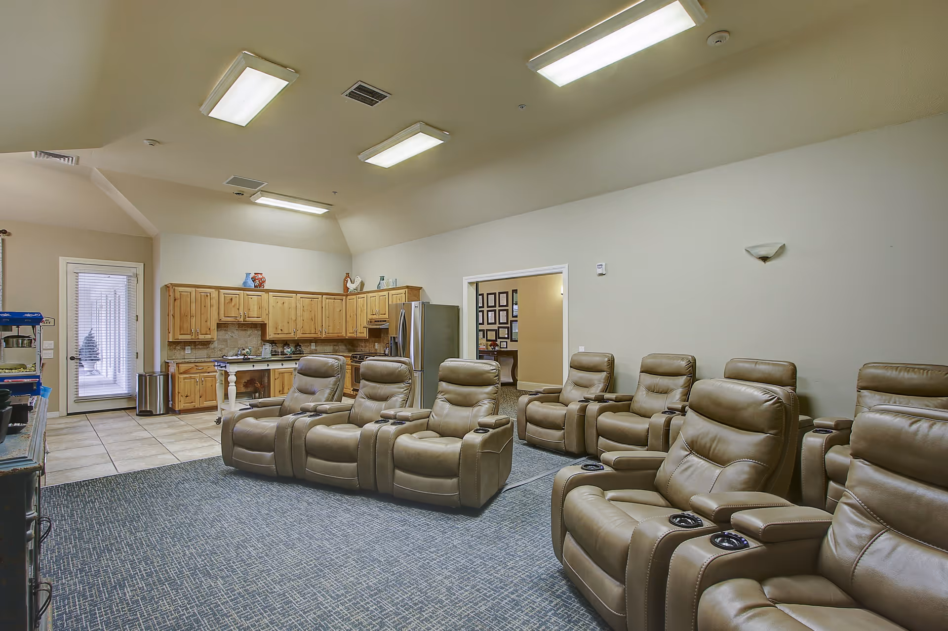 Interior view of a senior living facility common area with a row of beige leather recliner chairs arranged on a carpeted floor. In the background, there is a kitchenette with wooden cabinets, a stainless steel refrigerator, and a door leading outside. The room is well-lit with ceiling lights.