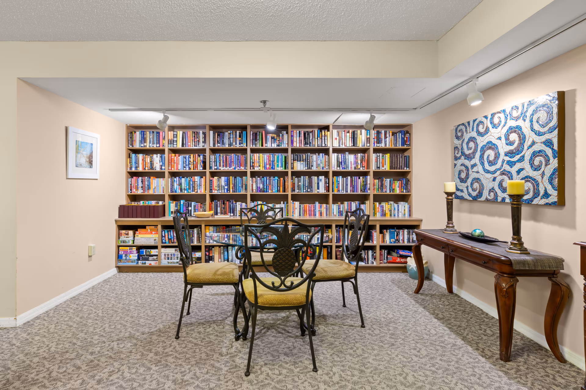 A cozy reading room with a large wooden bookshelf filled with books and board games. In front of the bookshelf is a round glass table with four ornate metal chairs with yellow cushions. To the right, there is a wooden console table with two candle holders and decorative items, and above it hangs a colorful abstract artwork with swirling patterns. The room has beige walls and patterned carpet flooring.