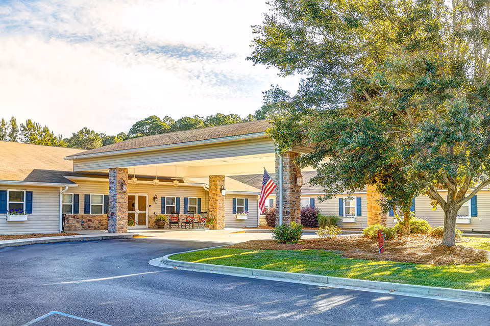 Front entrance of a single-story senior living building with a covered porte-cochère, American flag, and landscaped grounds.