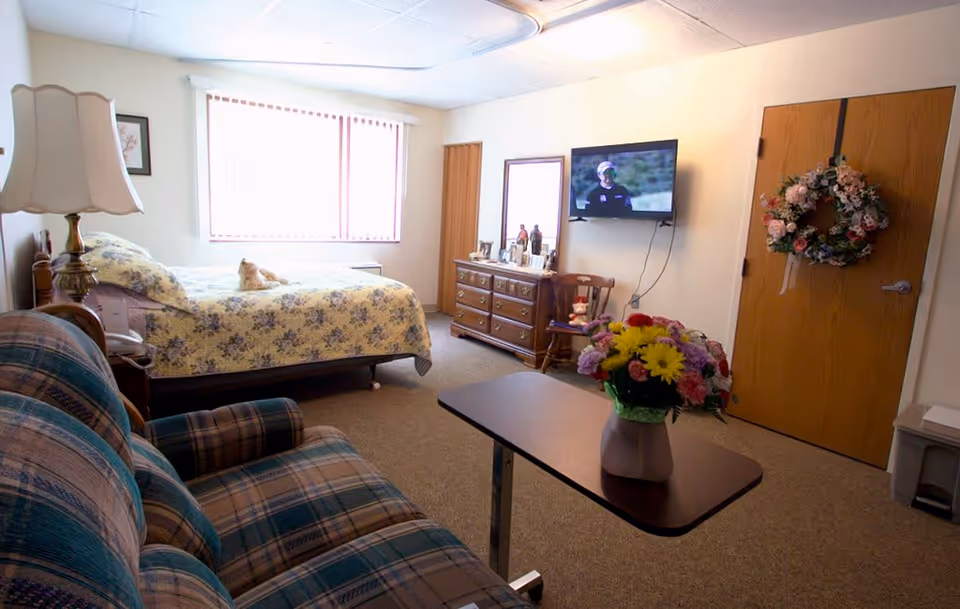 A cozy bedroom in St. Andrew's Village featuring a bed with floral bedding, a plaid upholstered couch, a wooden dresser with framed photos, a wall-mounted TV, a wooden door decorated with a floral wreath, and a table with a vase of colorful flowers.