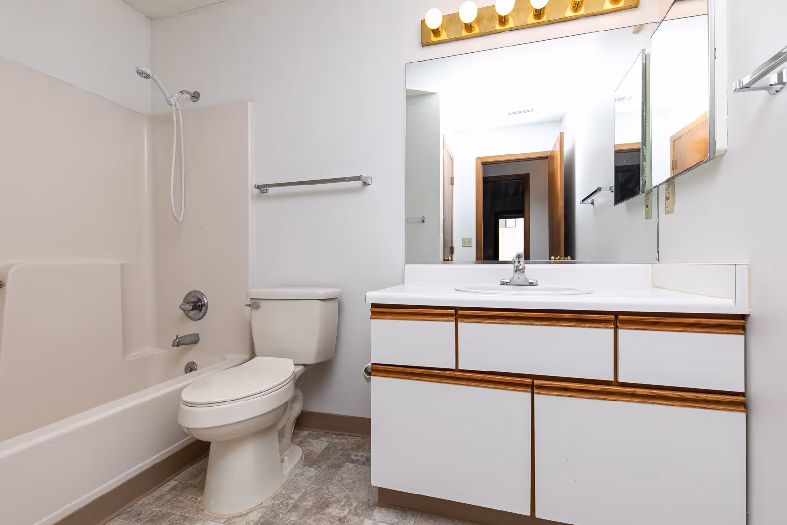 A clean bathroom featuring a beige bathtub with a showerhead, a white toilet, and a white vanity with wooden trim. Above the vanity is a large mirror with a row of lights mounted on a gold fixture. The floor has a patterned tile design, and there is a towel rack mounted on the wall next to the toilet.