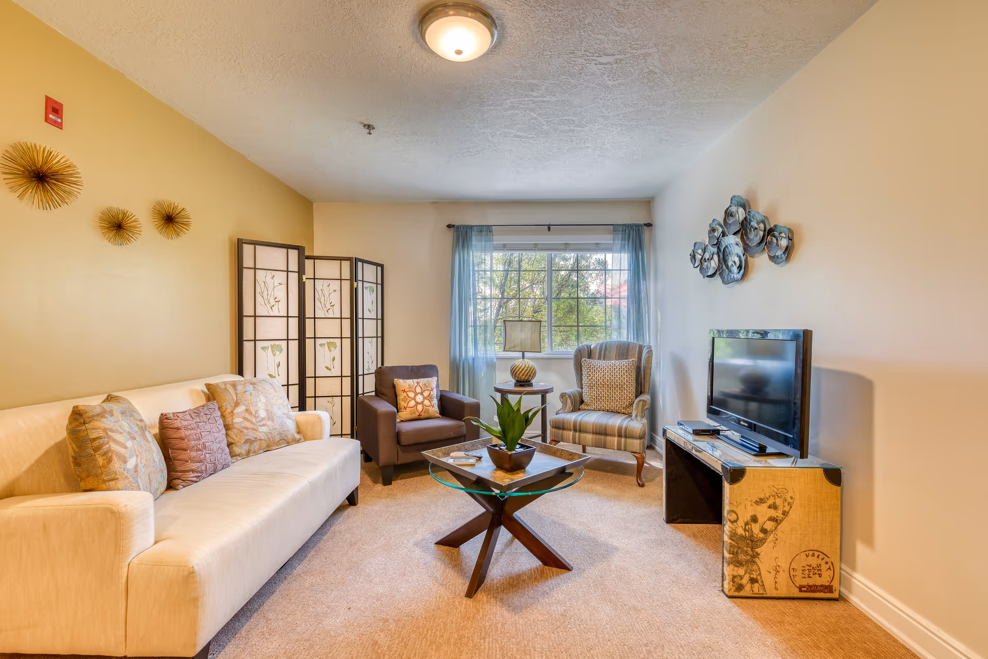 A cozy living room with a beige sofa adorned with decorative pillows, a brown armchair, and a striped armchair. A glass-top coffee table with a plant sits in the center. A TV is placed on a vintage-style console against the wall. The room has a window with sheer blue curtains letting in natural light, and decorative wall art is displayed on the walls.
