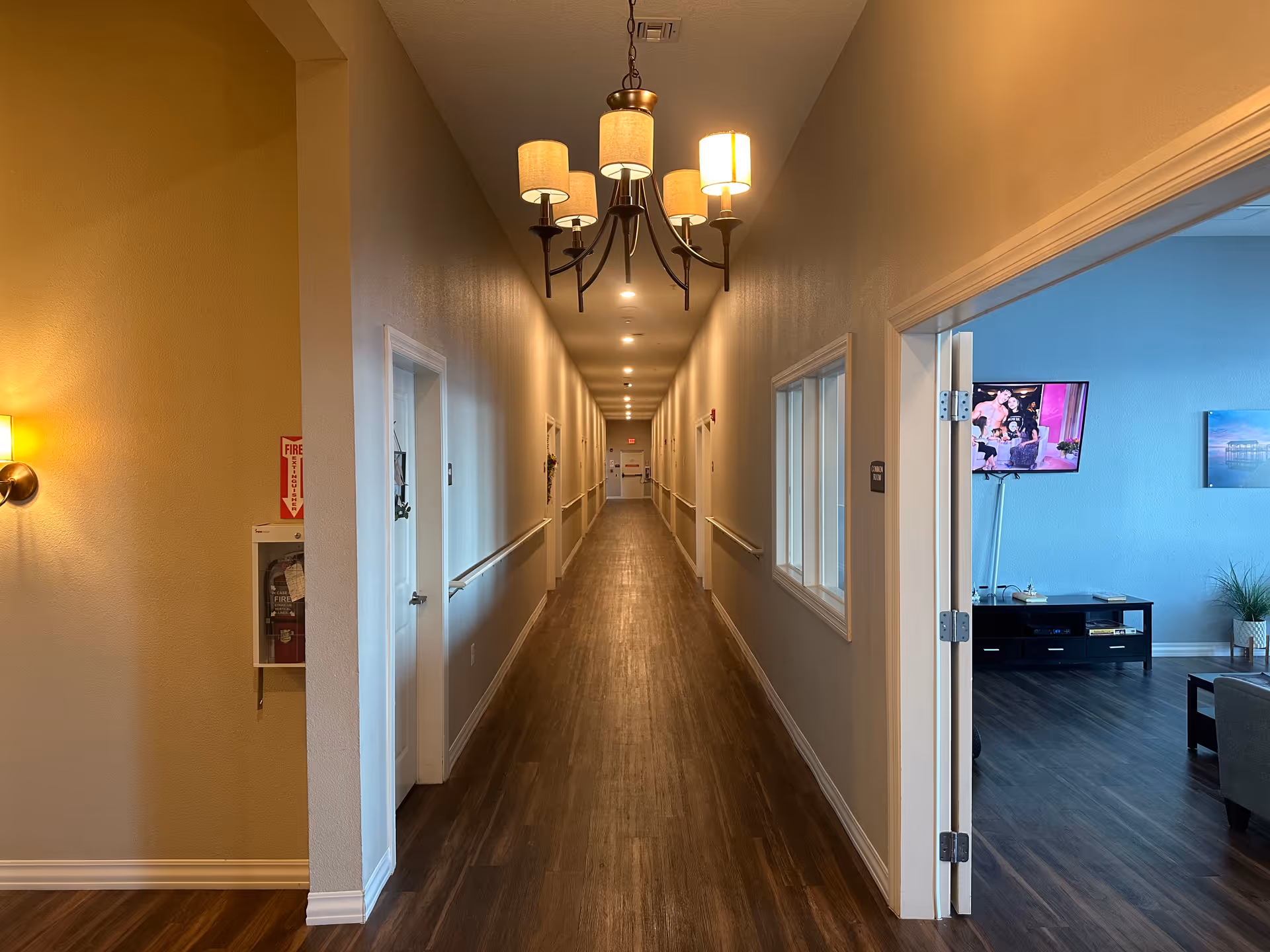 Long hallway in an assisted living facility with wooden flooring, beige walls, and ceiling lights. On the left side, there is a fire extinguisher cabinet and a wall sconce light. On the right side, an open door reveals a room with blue walls, a TV mounted on a stand, a couch, and a framed picture on the wall.