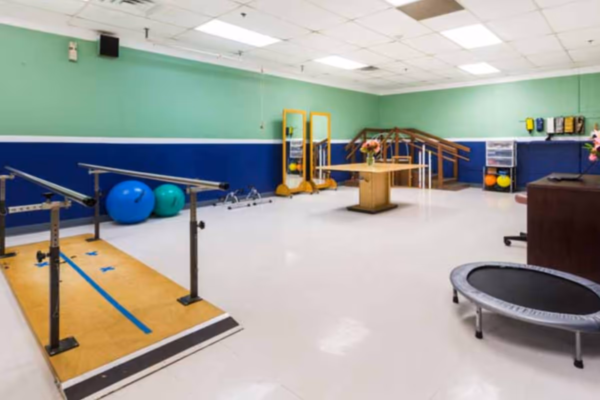 A physical therapy or rehabilitation room with parallel bars for walking exercises, exercise balls, a small trampoline, a wooden table with a flower vase, and a set of stairs with handrails for mobility training. The walls are painted green and blue, and the floor is white.