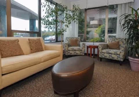 A cozy seating area with a beige sofa, two patterned armchairs, a dark brown oval ottoman, and a small wooden side table. Large windows let in natural light and show some greenery outside. There are also two potted plants inside the room.