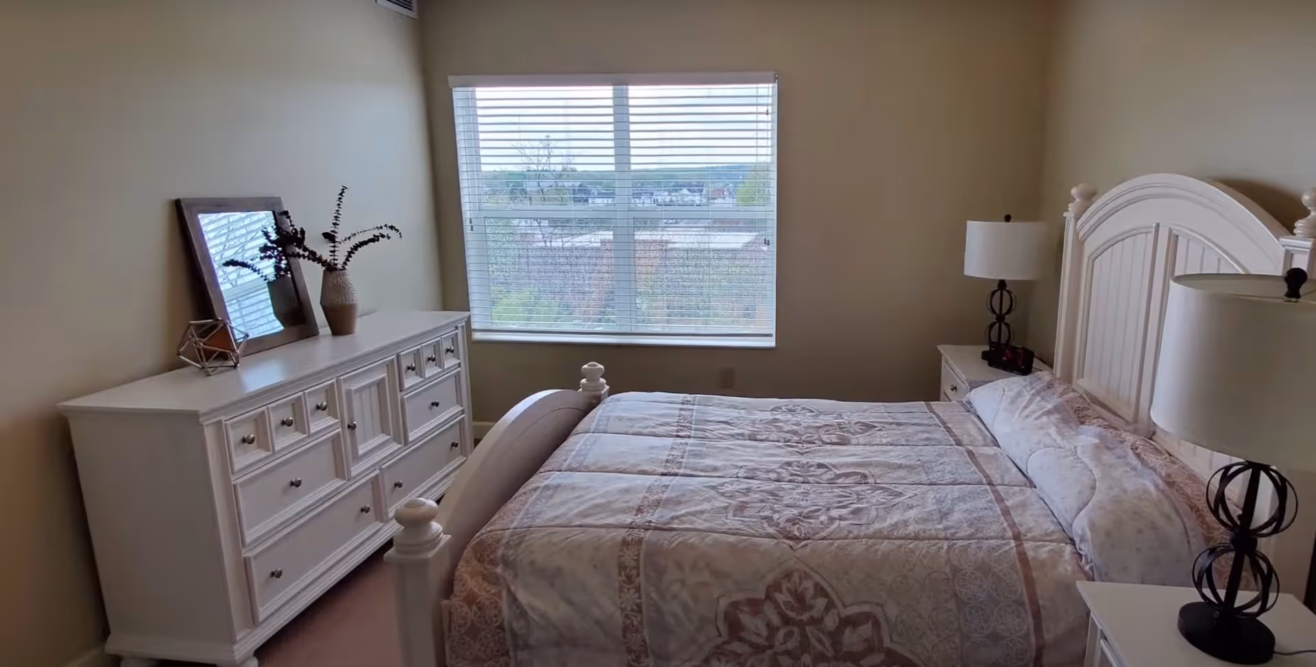 A bedroom with a large window covered by white blinds. The room features a white wooden bed with a patterned bedspread, two bedside tables each with a lamp, and a white dresser with a mirror and decorative items on top. The walls are painted beige.