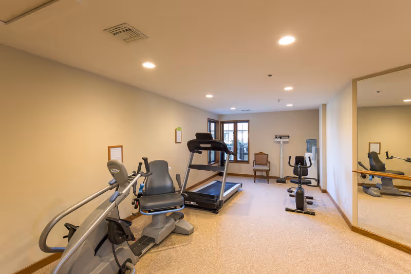 A small fitness room with exercise equipment including a recumbent bike, treadmill, and stationary bike. There is a chair and a scale near the window at the far end of the room. The room has beige walls, carpeted floor, recessed ceiling lights, and a large mirror on the right wall.