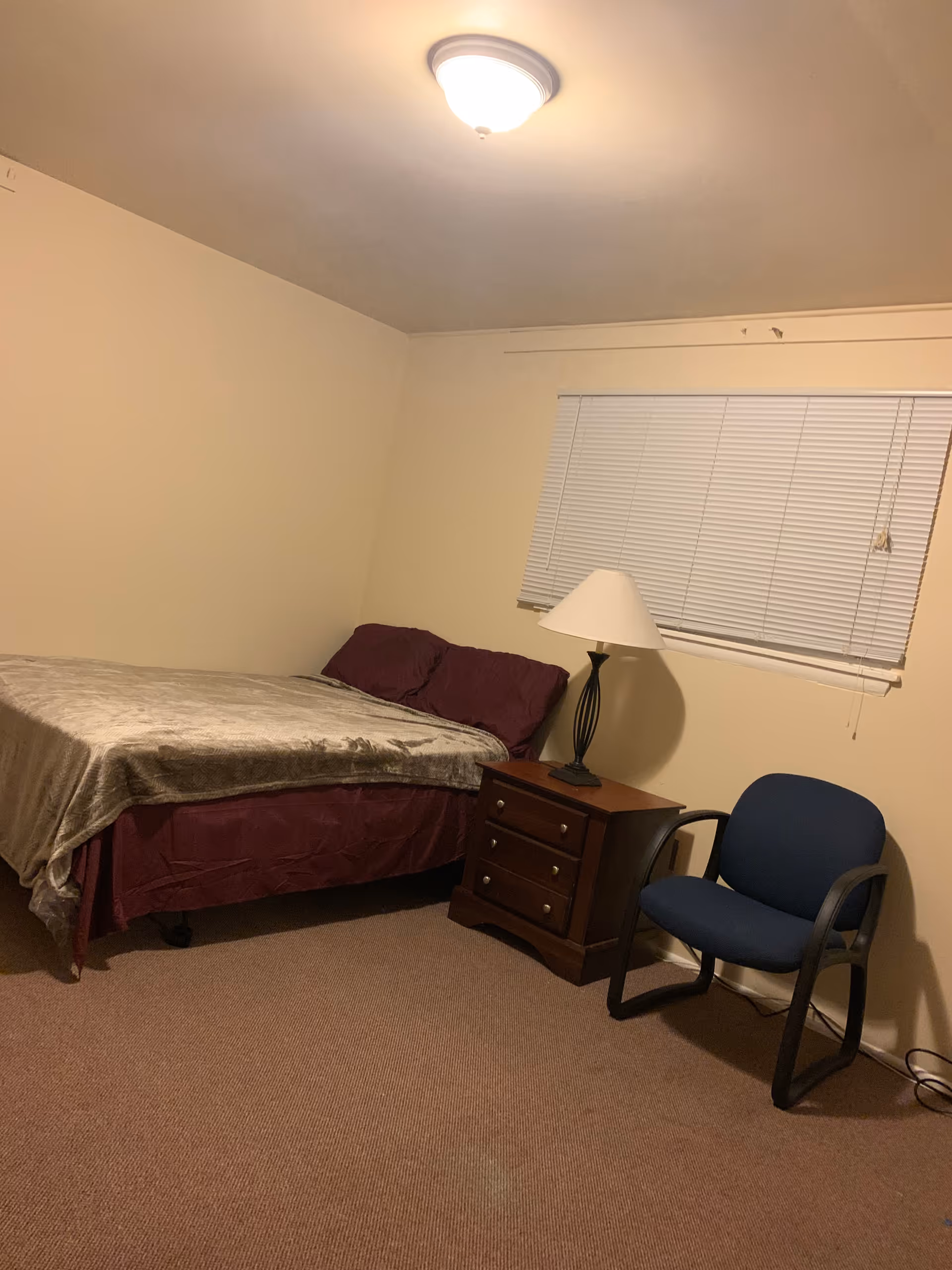A simple bedroom with a bed covered in a gray blanket and dark red sheets, a wooden nightstand with a lamp, a blue cushioned chair, beige walls, a window with closed blinds, and a ceiling light fixture.