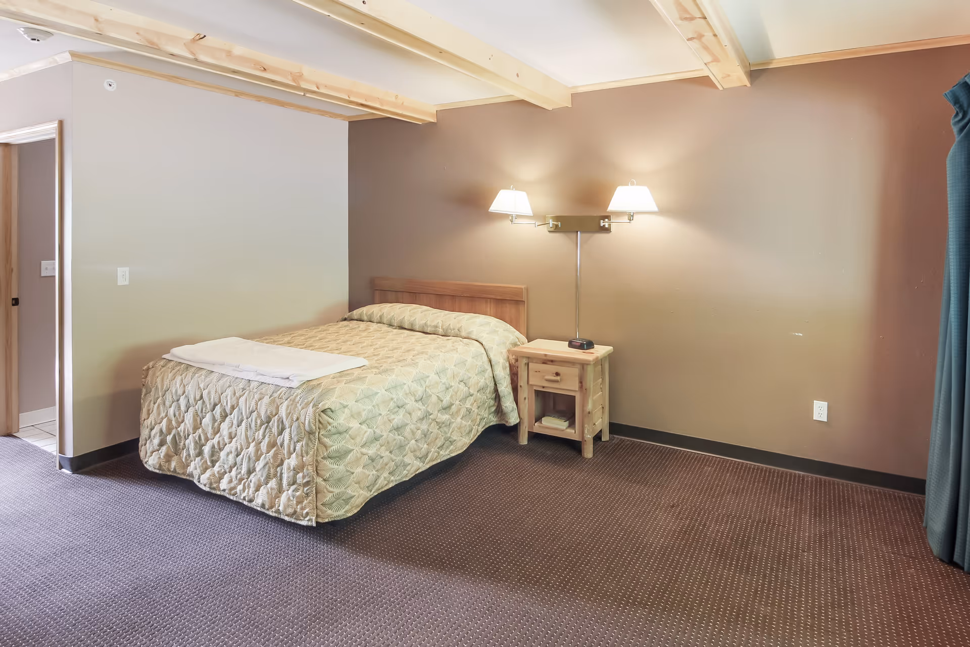 A simple bedroom with a bed covered in a patterned beige quilt, a wooden nightstand with a drawer and shelf, and a wall-mounted double lamp above the nightstand. The room has brown carpet, beige and brown walls, exposed wooden ceiling beams, and a partially visible doorway and curtain.