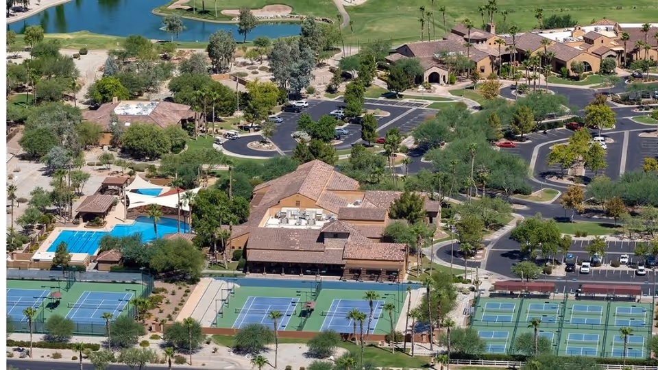 Aerial view of a community with a clubhouse, swimming pool, and multiple tennis courts surrounded by homes, parking areas, and landscaping.