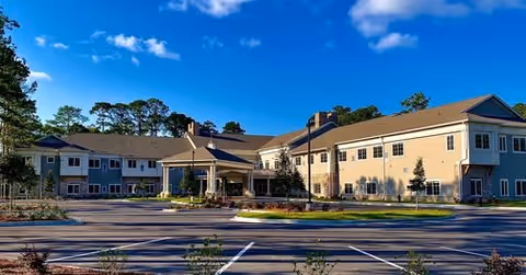 Exterior view of a two-story senior living facility building under a clear blue sky with a parking lot in the foreground and landscaped greenery around the building.