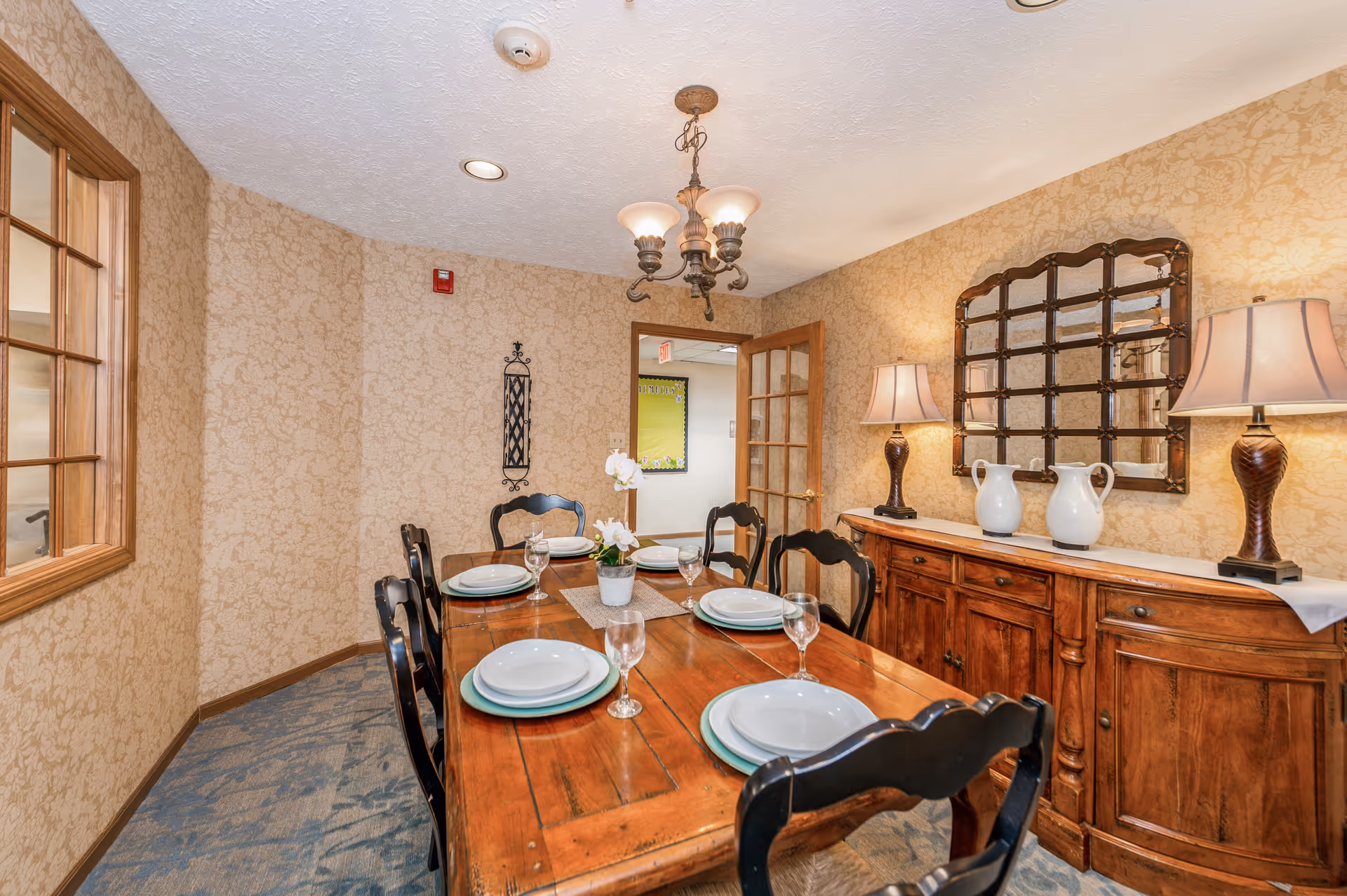 A dining room with a wooden table set for six people with white plates and glasses. The room has beige patterned wallpaper, a wooden sideboard with two lamps and two white pitchers, and a decorative mirror above the sideboard. There is a chandelier hanging from the ceiling and a door with glass panels leading to another room.