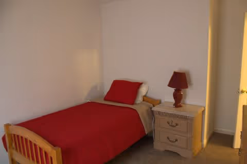A small bedroom with a single wooden bed covered with a red blanket and a white pillow. Next to the bed is a beige nightstand with two drawers and a red lamp on top. The room has plain white walls and an open door leading to another area.