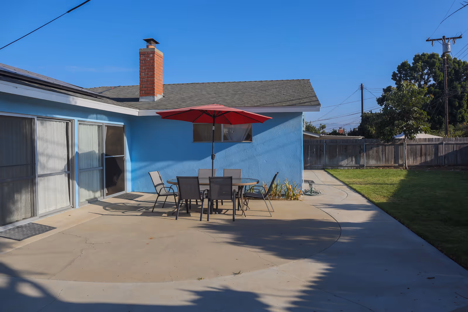Outdoor patio area with a round glass table and six chairs under a red umbrella, adjacent to a blue building with a brick chimney. The patio is surrounded by a concrete walkway and a grassy lawn with a wooden fence and trees in the background under a clear blue sky.