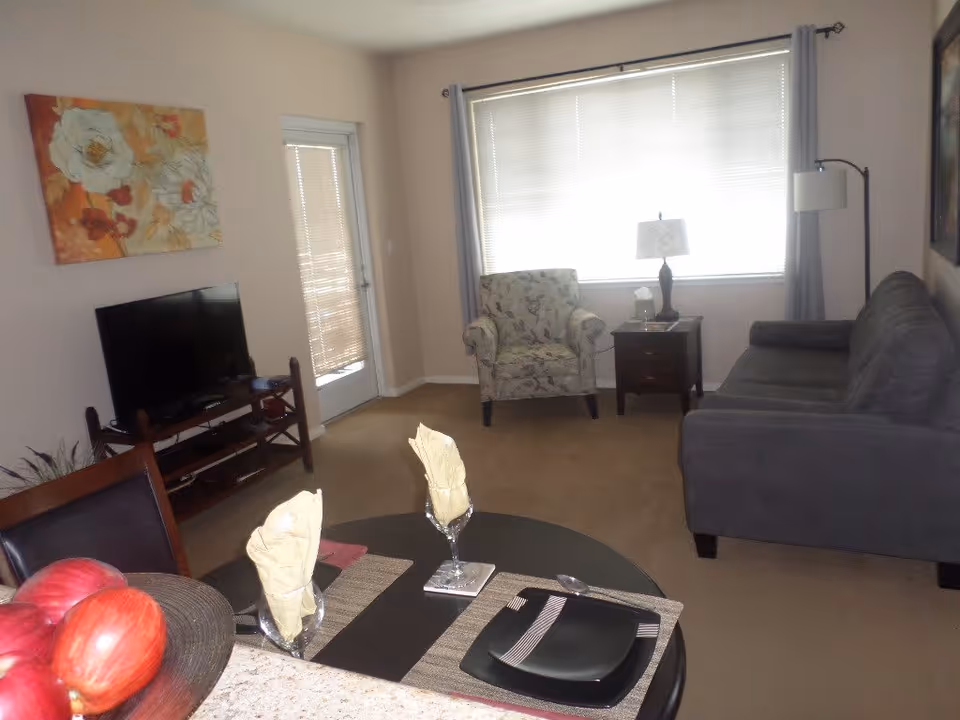 A cozy living room with a gray sofa, a floral armchair, a side table with a lamp, a TV on a wooden stand, and a round dining table set with black plates and napkins. There is a large window with blinds and curtains, and a door with blinds next to the TV. A floral painting hangs on the wall above the TV.