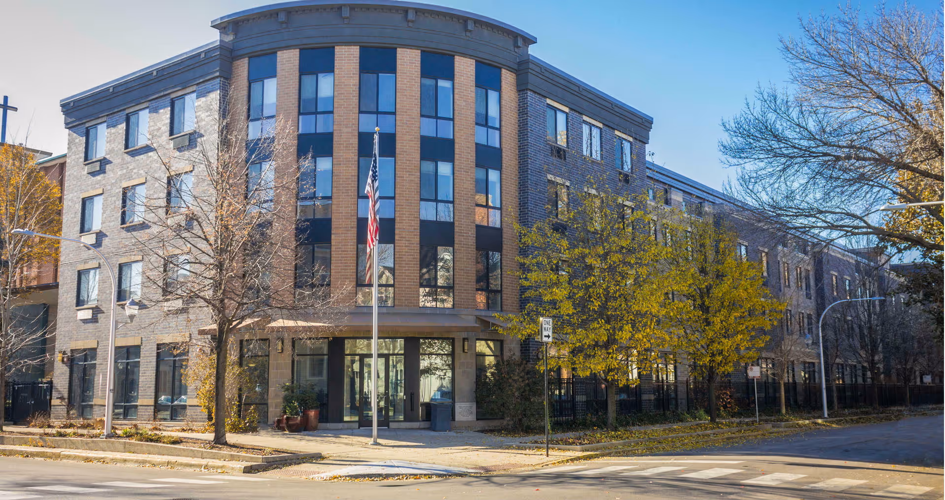 Exterior view of a four-story senior living facility building with large windows and a central entrance. There is an American flag on a flagpole in front of the building, surrounded by trees with autumn foliage. The sky is clear and blue.