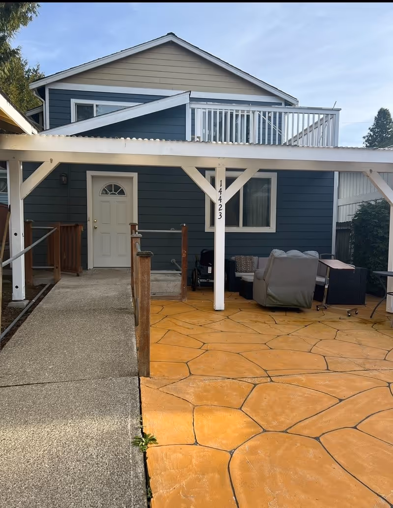 Exterior view of a two-story house with blue siding and white trim. The house has a white door, a window, and a balcony with white railing on the second floor. There is a covered patio area with a white pergola structure, a concrete ramp with wooden handrails leading to the door, and orange stone-patterned flooring. Outdoor furniture including a sofa and a table are placed under the pergola. The house number 14423 is displayed on one of the pergola posts.