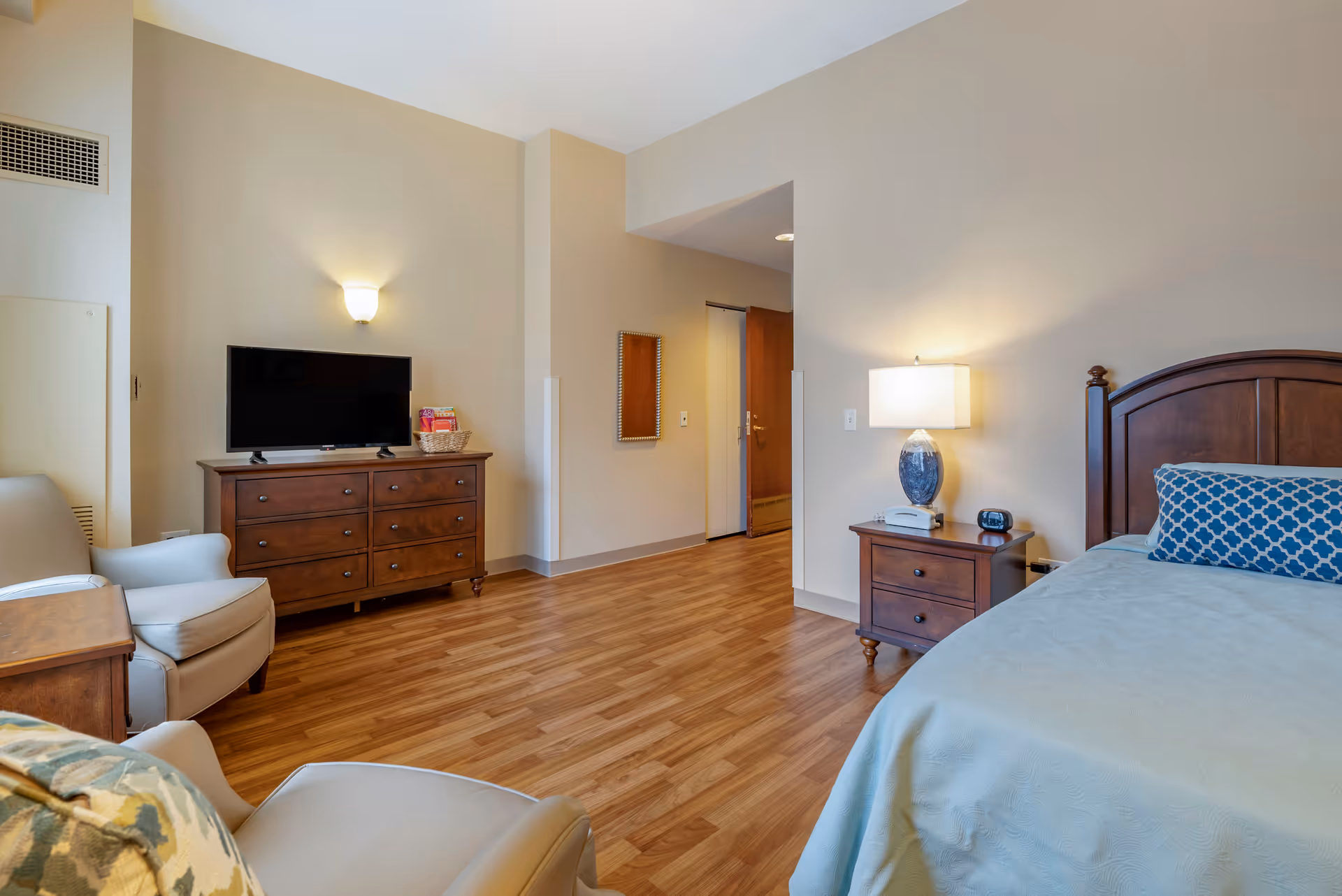 A senior living facility bedroom with a wooden bed featuring a blue patterned pillow and light blue bedspread. There is a wooden nightstand with a lamp, telephone, and clock. Across the room is a wooden dresser with a flat-screen TV and a basket with magazines. Two beige armchairs and a small wooden table are also visible. The room has wood flooring and beige walls with a wall-mounted light fixture.