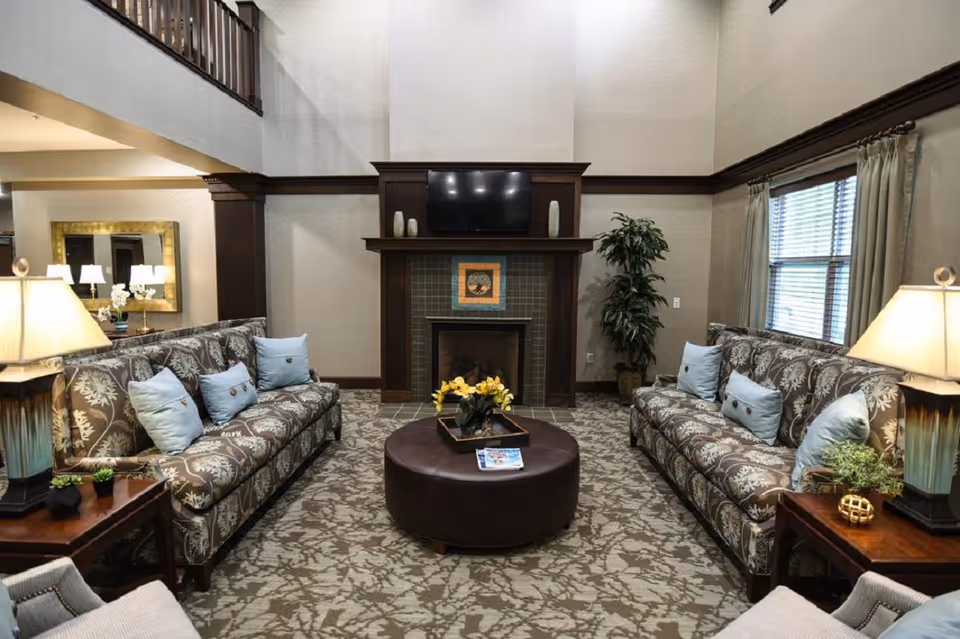 A cozy living room with two patterned sofas facing each other, each adorned with light blue pillows. Between the sofas is a round brown ottoman with a tray holding a yellow flower arrangement and some magazines. The room features a fireplace with a mounted flat-screen TV above it, flanked by decorative vases. There are two side tables with lamps on either side of the sofas, a large window with curtains on the right, and a tall plant next to the fireplace. The walls are light-colored with dark wood trim, and the carpet has a floral pattern.