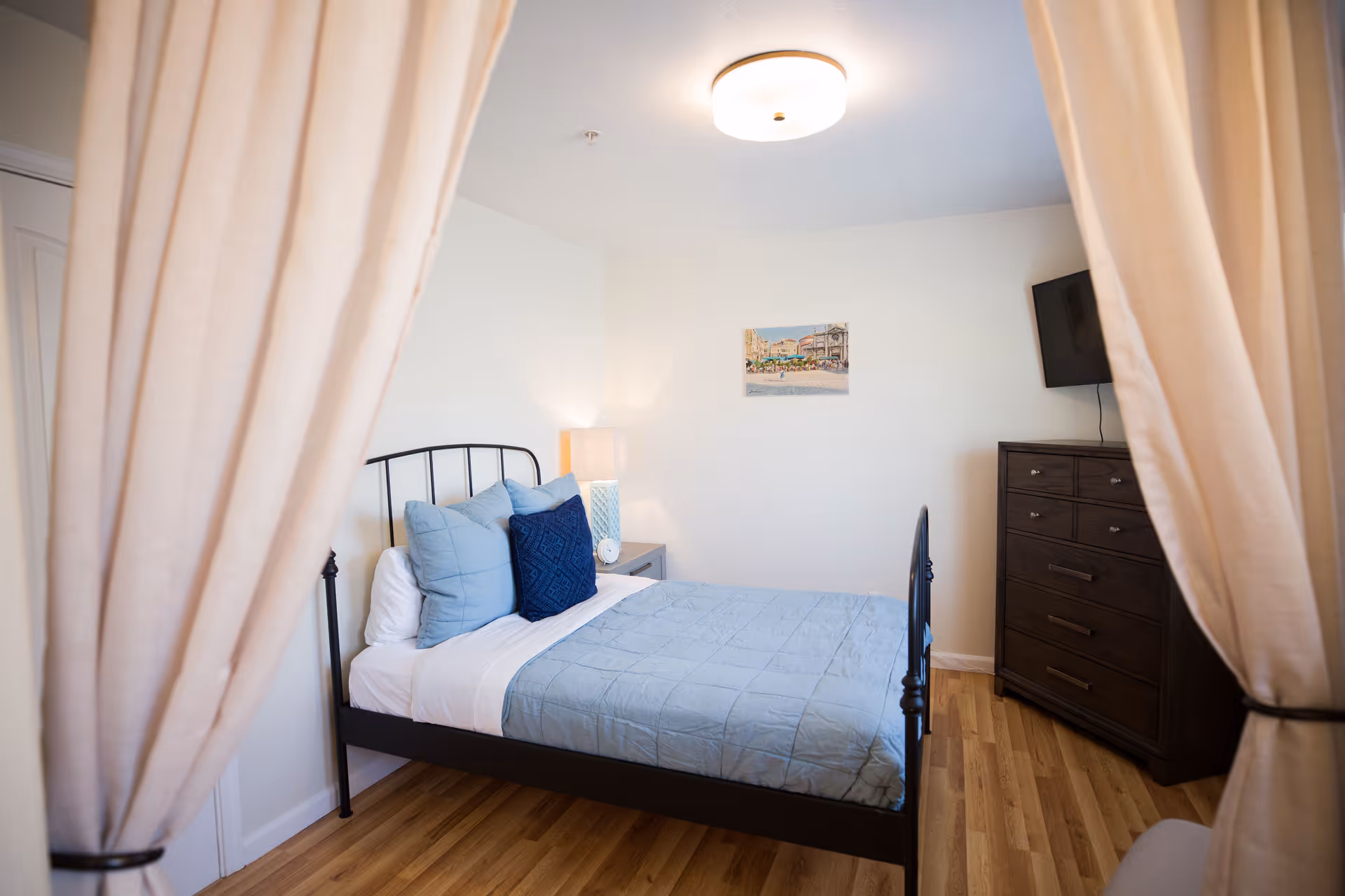 A cozy bedroom with a black metal bed frame, light blue bedding, and several pillows. There is a nightstand with a lamp and an alarm clock next to the bed. A dark wooden dresser with a mounted flat-screen TV is against the wall. The room has light-colored walls, wood flooring, and is partially framed by beige curtains.