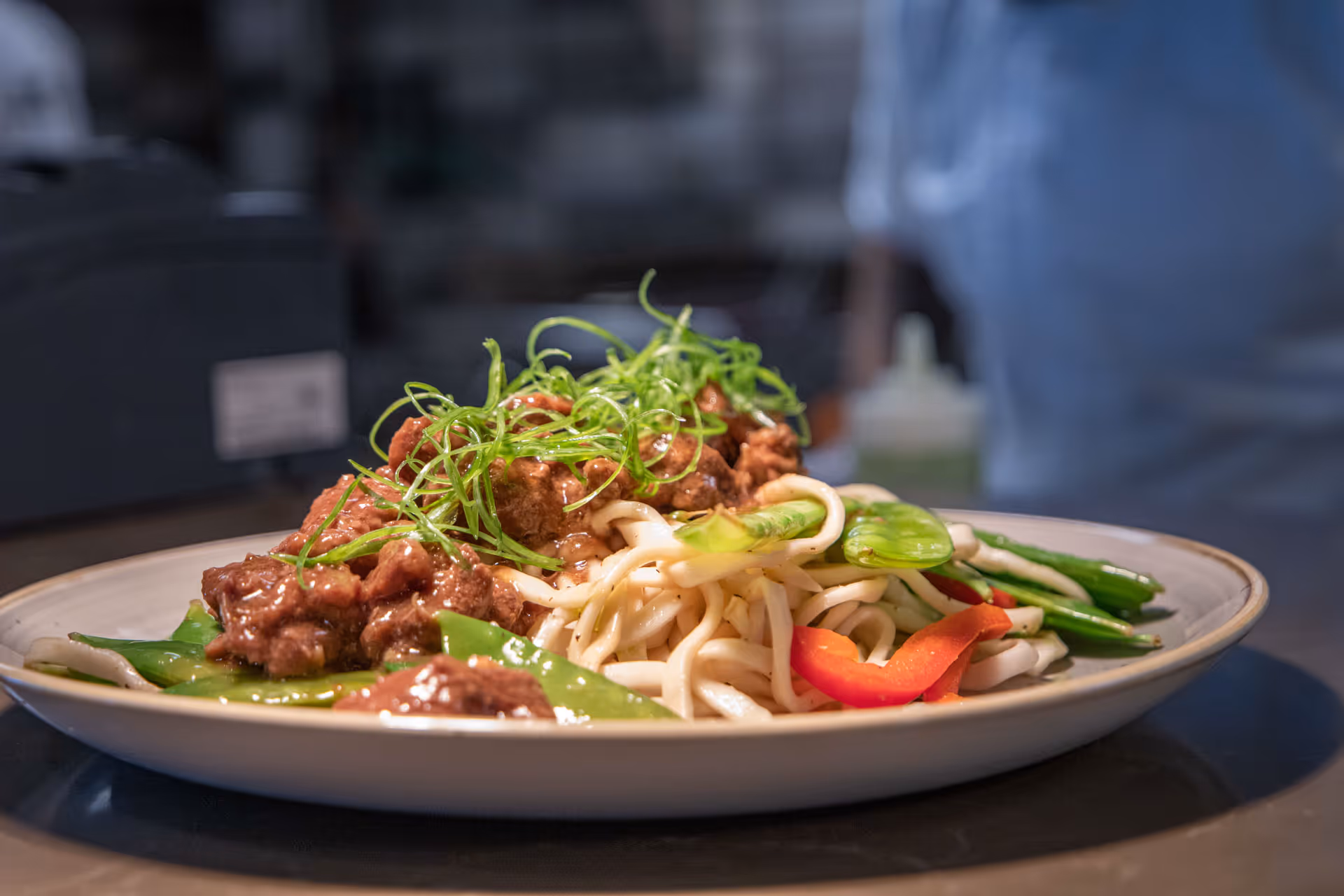 A plate of noodles topped with cooked beef, green vegetables, and garnished with thinly sliced green onions, served on a white plate.