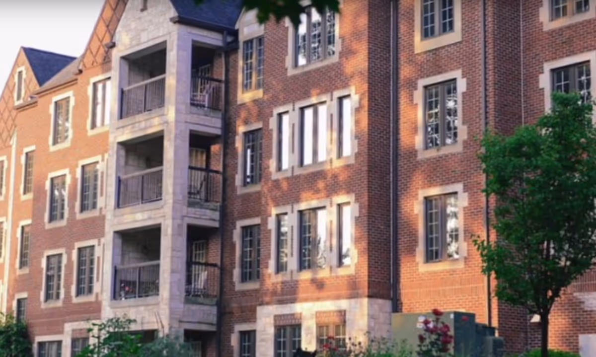 Exterior view of a multi-story brick building with multiple windows and balconies, surrounded by greenery and trees, taken during daylight.