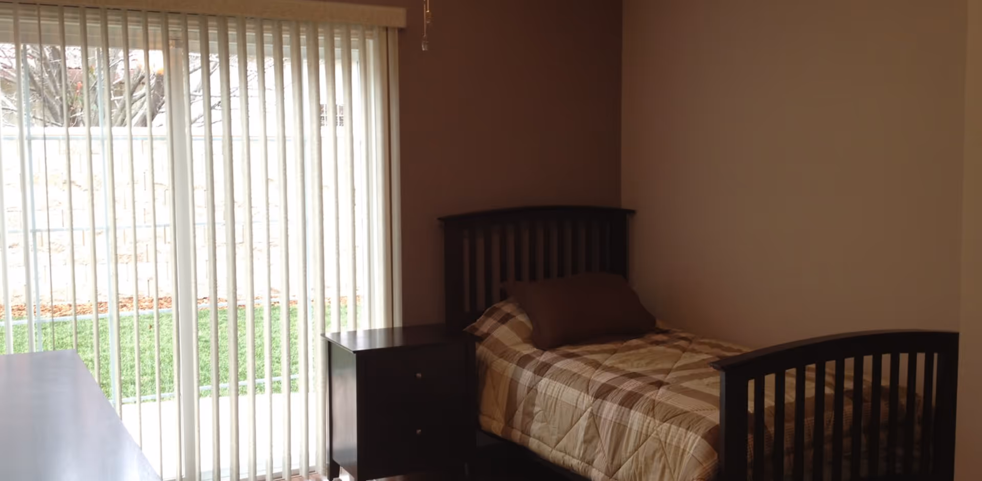 Small bedroom with a twin bed and nightstand beside a sliding glass door with vertical blinds looking out onto a lawn.