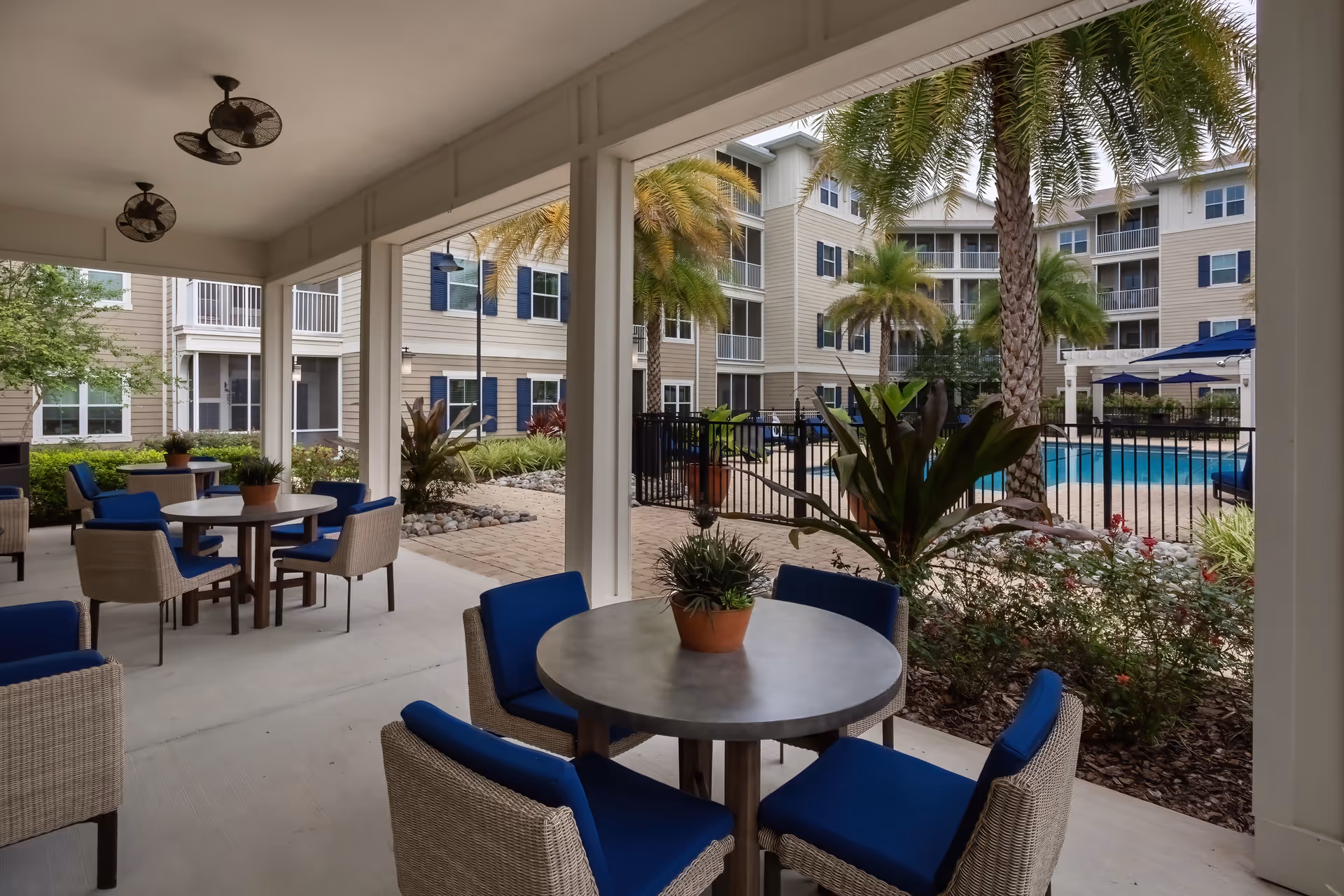 Covered outdoor seating area with round tables and wicker chairs with blue cushions, overlooking a fenced swimming pool surrounded by palm trees and a multi-story residential building in the background.