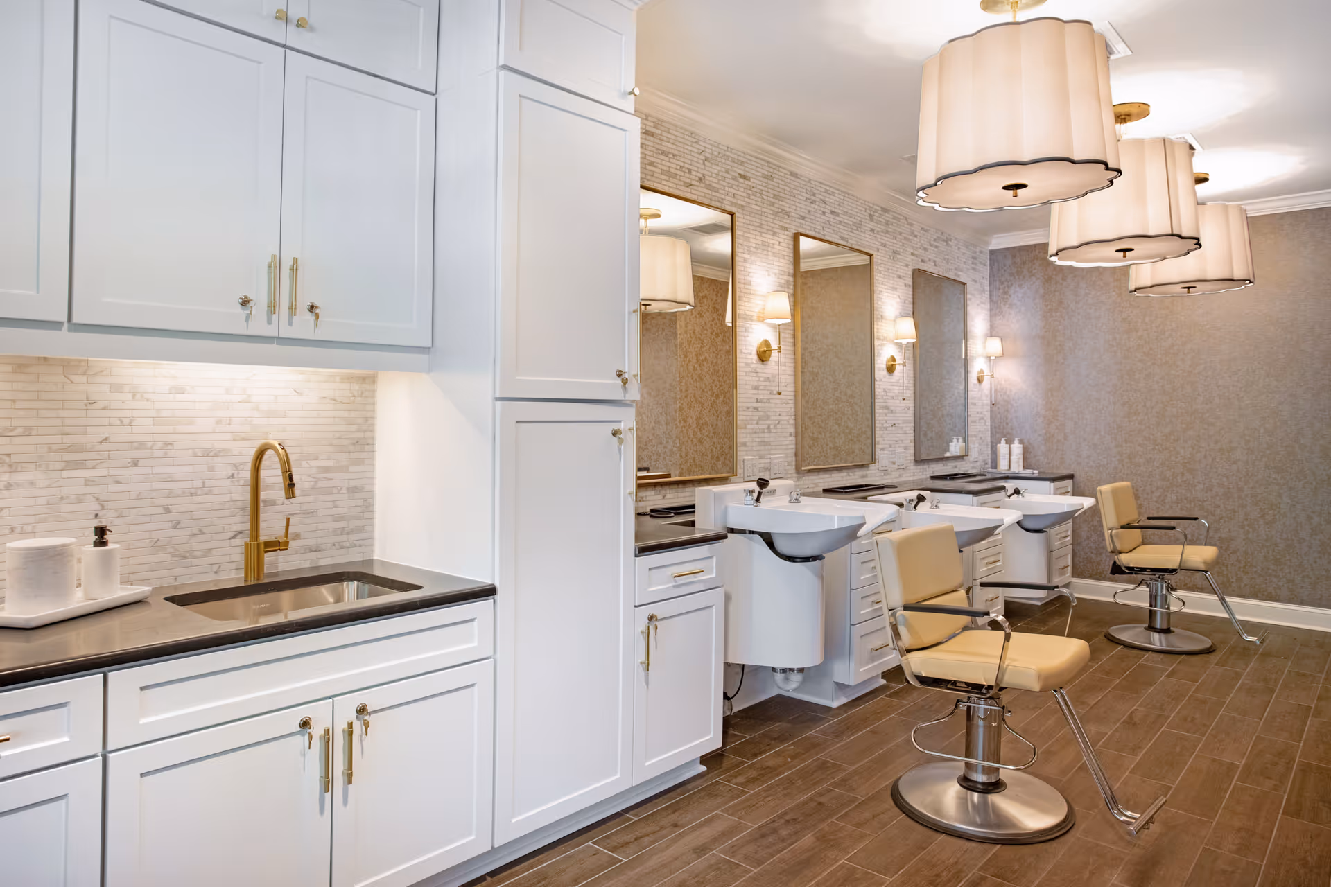Interior view of a salon area in a senior living facility featuring three beige salon chairs in front of three white wash basins with mirrors above. The room has white cabinetry with gold handles, a black countertop with a sink and gold faucet, and warm lighting from wall sconces and large ceiling lamps. The floor is covered with brown tiles and the walls have a textured wallpaper.