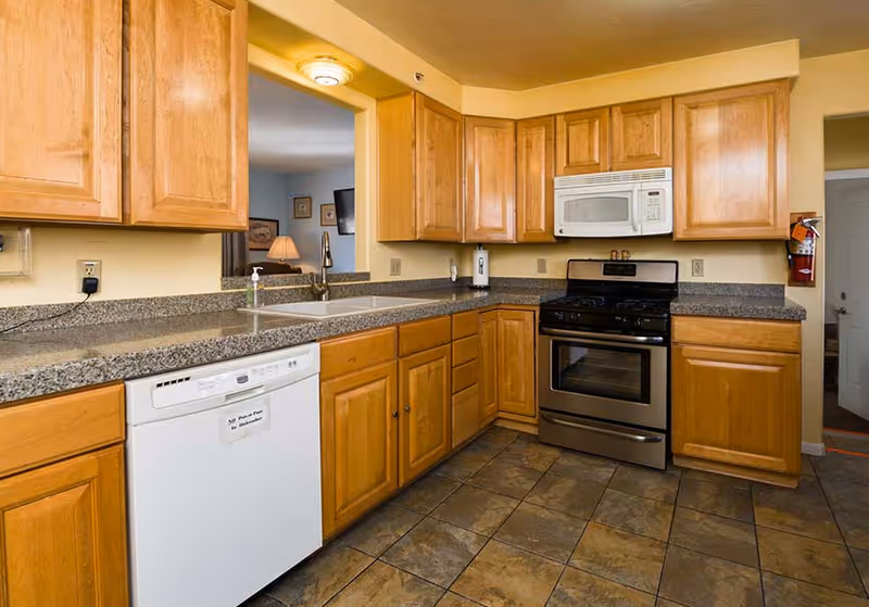 A clean kitchen with wooden cabinets, granite countertops, a white dishwasher, a double sink, a stainless steel gas stove with an oven, and a white microwave above the stove. The floor is tiled, and there is a fire extinguisher mounted on the wall near a doorway.