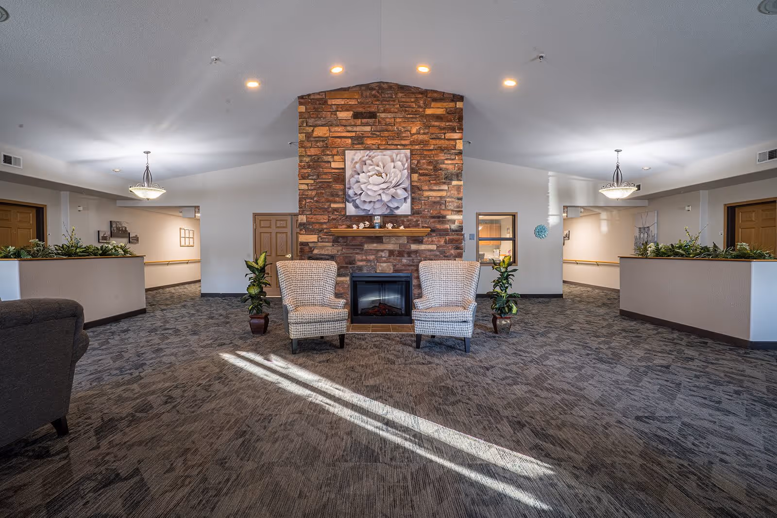 A spacious common area with a stone fireplace in the center, flanked by two patterned armchairs and two potted plants. The room has carpeted flooring, white walls, and recessed lighting. There are doorways and windows visible on either side of the fireplace, with decorative plants on half walls and ceiling light fixtures.