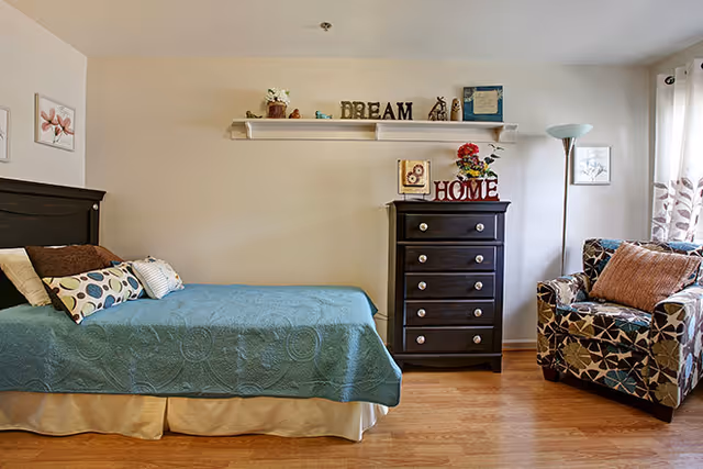 A cozy bedroom featuring a single bed with a teal coverlet, a patterned armchair, a dark chest of drawers topped with decorative 'HOME' letters, and a wall shelf displaying 'DREAM'.