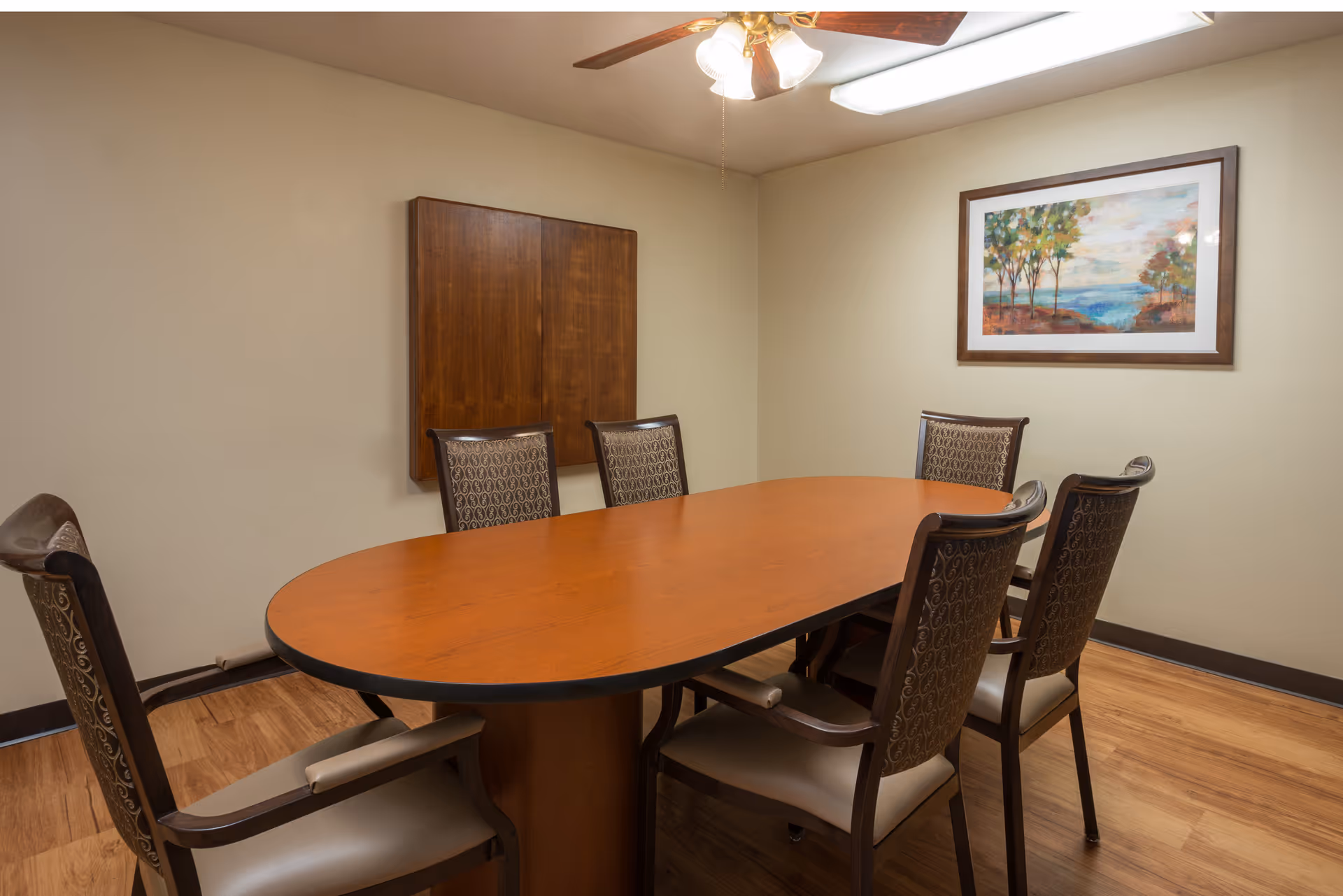 A small meeting or dining room with a wooden oval table surrounded by six cushioned chairs. The room has beige walls, a ceiling fan with lights, a wooden floor, a framed landscape painting on the wall, and a closed wooden cabinet.