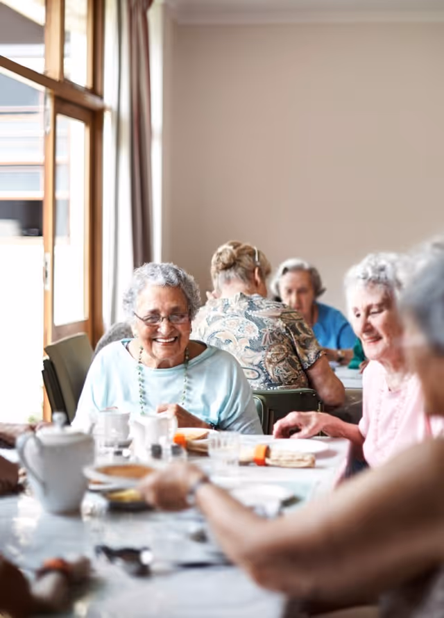 A group of elderly women sitting around a table enjoying a meal together in a bright room with large windows and curtains.